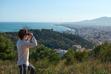 A shallow focus shot of a young man from Spain in a white shirt wearing a mask observing the coastal town through binoculars