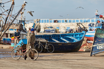 Man at fish market in Essaouira/Morocco pushing his bike and rubbing his eyes. Empty boat lies...