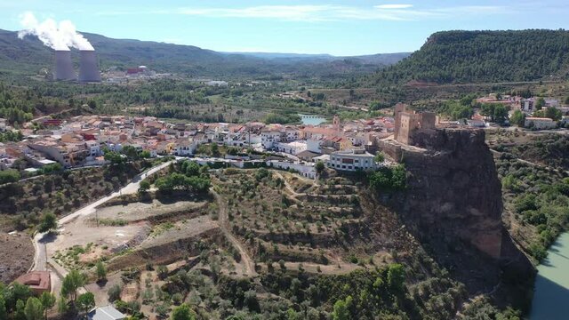 Aerial view of historical Spanish town of Cofrentes with medieval castle on top of basalt rock and modern nuclear power plant cooling towers on background, Valencia, Spain