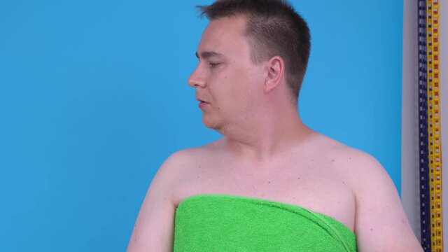 Young man wrapped in green bath towel cringes in disgust, then changes his mood, begins to smile and shows thumbs-up gesture of approval, blue background, studio shooting. Actor on audition.