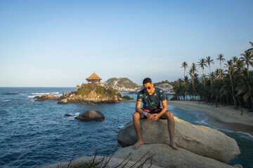 Fototapeta na wymiar Hombre joven con gafas de vacaciones sentado en una roca junto al mar azul en un día soleado de verano 