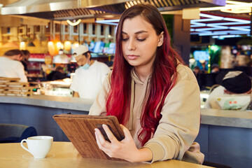 A lovely woman with red hair sits in a cafe, works on a tablet, in a restaurant with a cup of coffee.