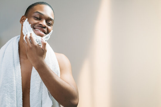Male skincare. Cosmetic product advertising. Grooming hygiene. Happy shirtless African man with white towel enjoying applying shaving foam on face skin isolated on light copy space background.