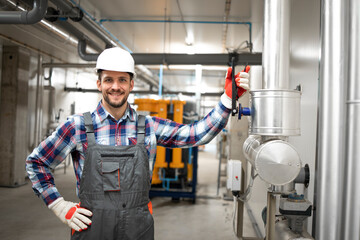 Portrait of factory engineer worker holding his arm on the valve and standing by heating or cooling pipeline system at boiler room. Gas installation maintenance.