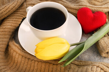 A white cup of coffee stands next to a yellow tulip with a red heart and a knitted sweater.
