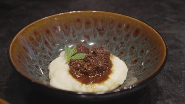 Chefs makes plating of meat and mashed potato dish, adding bits of green veggies nn top. Close up, slow motion shot.