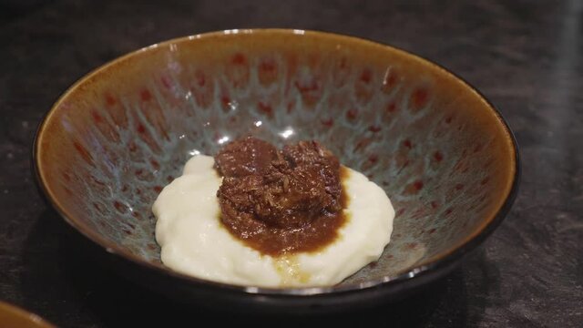 Chefs makes plating of meat and mashed potato dish, adding bits of green veggies nn top. Close up, slow motion shot.