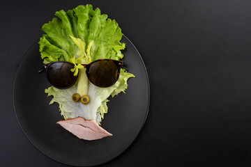 creative concept, cubist style portrait made from lettuce, apio, pastrami and olives, with sun glasses, isolated on a black ceramic plate