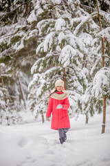 A 10-11-year-old girl walks through a snow-covered park. She's wearing a white knit hat and scarf. She's wearing a pink down jacket. She's having fun, throwing snow. She walks with a teddy bear.