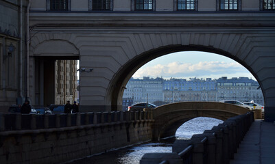 Winter Canal in St. Petersburg, arch over it, on the opposite bank of the Neva river old houses, bright spring sky with clouds, cars pass over the bridge, pedestrians walk. bridge, river, architecture