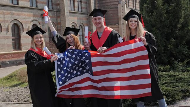 A group of American university graduates in robes, hats with diplomas on graduation from the academy hold in their hands the American Flag laughing dancing and posing for the camera academy background