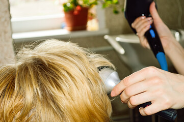 close-up - hairdresser dries hair with a hairdryer and styles with a round comb short, blonde hair of a woman after a haircut