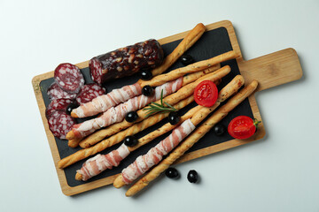 Board with grissini sticks with bacon and snacks on white background