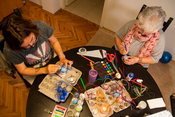 Easter egg painting and decorating activity concept: Cheerful mother with white hair and adult daughter enjoying togetherness and creative indoor decoration. Handmade colorful funny eggs