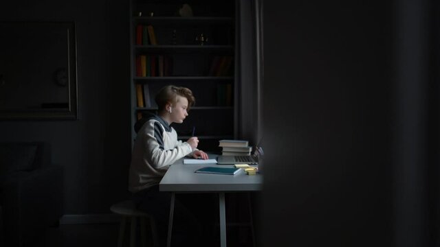 Side view of pupil boy wearing wireless earphone learning online using laptop making notes in copybook sitting at desk at home near window. Concept of distance education. Tracking shot in slow motion
