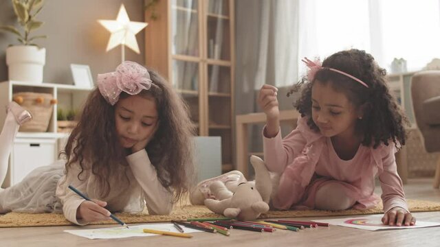 Steadicam of two curly Mixed-Race little sisters wearing cute headbands, lying on stomachs on floor of room at home, drawing and coloring with pencils