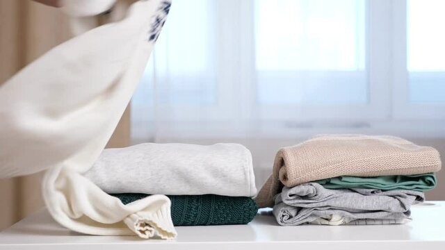Woman takes sweater from middle of folded knitwear clothes on white table against large window in brightly lit room extreme closeup. Lady looking for what to wear