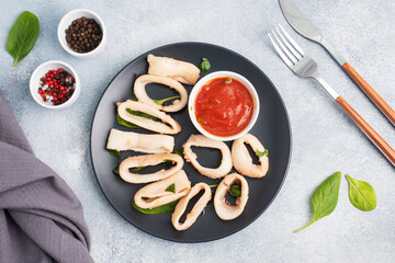 Fried squid rings with tomato sauce and lemon. Black plate, grey concrete background copy space. Top view