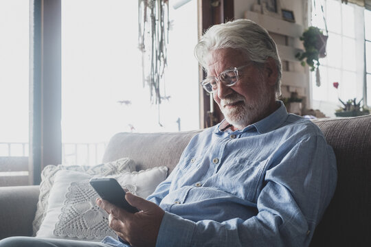 One old and mature man using phone, working with smartphone sitting on the sofa at home indoor concept and business man lifestyle. Male pensioner and retired person having fun relaxing on couch.