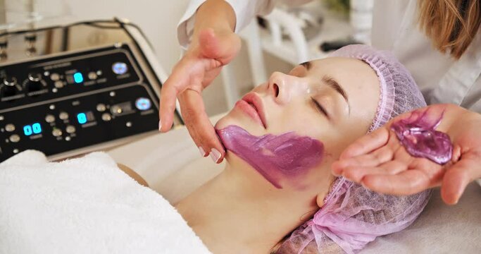 Cosmetologist applies a woman a therapeutic mask on her face. Woman in a spa salon on cosmetic procedures for facial care. Doctor does a massage of the girl's face with a moisturizing mask on a face.