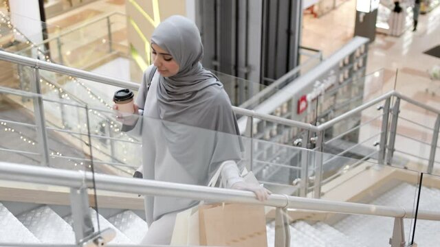 Lockdown of young attractive Muslim woman wearing grey abaya and hijab holding coffee paper cup and shopping bags going upstairs in mall during shopping