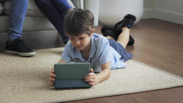 Cute little caucasian boy lying on floor carpet on stomach and playing and watching games on digital tablet at home in living room