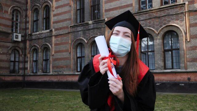 Young graduated redhead girl holding her graduation degree convocation ceremony. Attractive student graduate posing towards the camera dancing smiling in protective mask during coronavirus covid19.