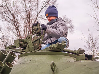 An eight-year-old Caucasian boy in a gray jacket and a blue knitted hat sits on a tank turret and is playing with the tower equipment.