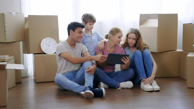 Cheerful and beautiful caucasian mother holding digital tablet and shopping for furniture for new home with handsome husband and children with cardboard boxes around