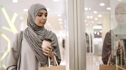Lockdown of young mixed-race woman wearing grey abaya and hijab going along mall with shopping bag and coffee paper cup in hands, stopping next to showcase and looking at it