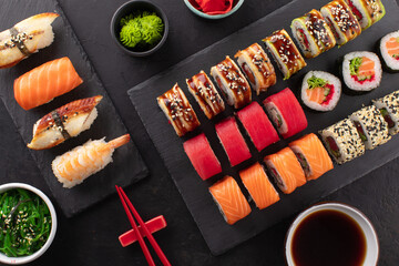Various kinds of sushi placed on black slate board, wakame chuka salad, bowl of soy sauce and ginger on the side. Asian food composition on black background, top view