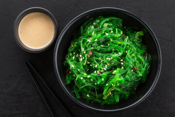 Healthy seaweed salad with sesame seeds in a black bowl and peanut sauce on the side on a black textured background. Top view.