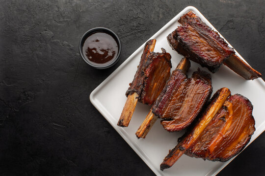 Smoked beef ribs with bone and barbecue sauce on a white square plate, top view, rustic black background. Roasted beef ribs with bone, copy space.