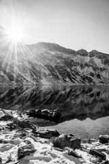 Black and white view of sunny mountainscape in Tatra National Park, Poland. Smooth water surface on lake, sunrays behind mountain ridge and fresh snow. Selective focus on the rocks, blurred background