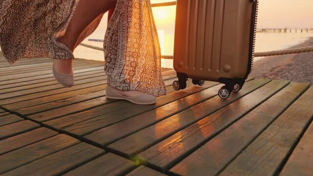 A tourist with a suitcase on the scroll came to the seaside resort and walks along the pier, close-up with a glare of the rising sun