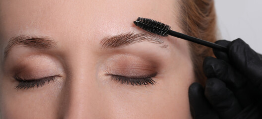 Beautician brushing woman's eyebrows before tinting on light grey background, closeup