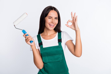 Photo of flirty lady hold painter roller show okey wink eye wear green overall isolated white color background