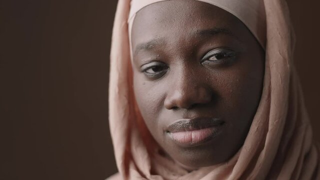 Close up of face of young Muslim African woman wearing pink hijab standing against brown background and looking at camera