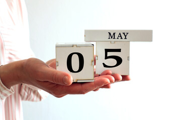 Calendar for May 5: cubes with the numbers 0 and 5, the name of the month in English in the hands of a young woman on a gray background, side view