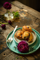 Three homemade cherry and chocolate muffins and ranunculus flowers on ceramic plates on old rustic wooden table. 