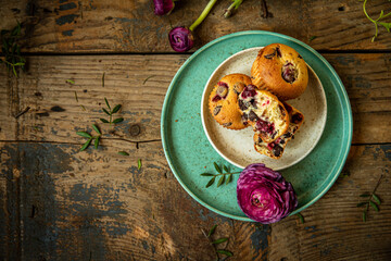 Three homemade cherry and chocolate muffins and ranunculus flowers on ceramic plates on old rustic wooden table. 