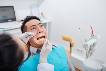 Scared young Asian man making funny face when getting his teeth treated in clinic