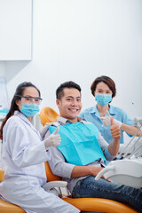 Happy young Asian patient, his dentist and assistant showing thumbs-up and smiling at camera after dental treatment