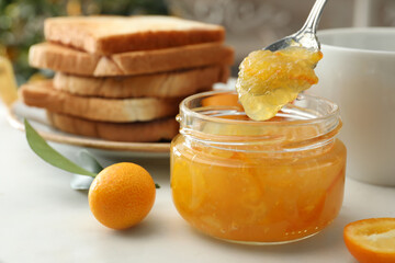 Spoon with tasty kumquat jam above jar on white table