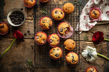 Group of homemade cherry and chocolate muffins on cooling rack and fresh flowers and leaves on rustic wooden table.