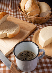 Breakfast table in Brazil with breads, cheese, cup of coffee and accessories on a brown and beige checkered tablecloth, dark background, selective focus.