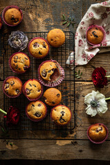 Group of homemade cherry and chocolate muffins on cooling rack and fresh flowers and leaves on rustic wooden table.