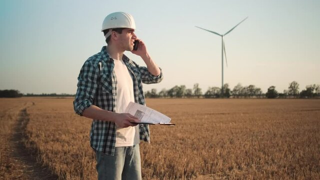 A side view of a worker man engineer in a hard hat is talking on his smartphone standing at the electric windmill farm