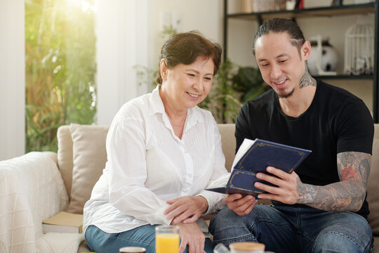Smiling mother and her adult son looking at old photos in album when spending time together on weekend