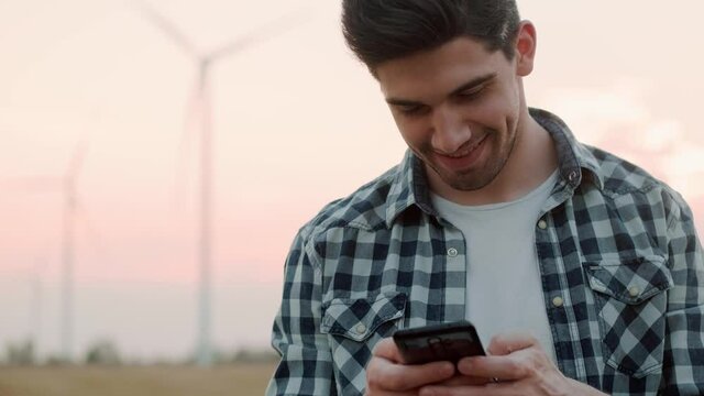 A happy man wearing shirt is using his smartphone standing at the electric windmill farm